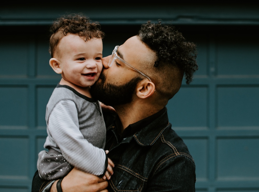 Father wearing glasses kissing one-year-old son in front of a blue background
